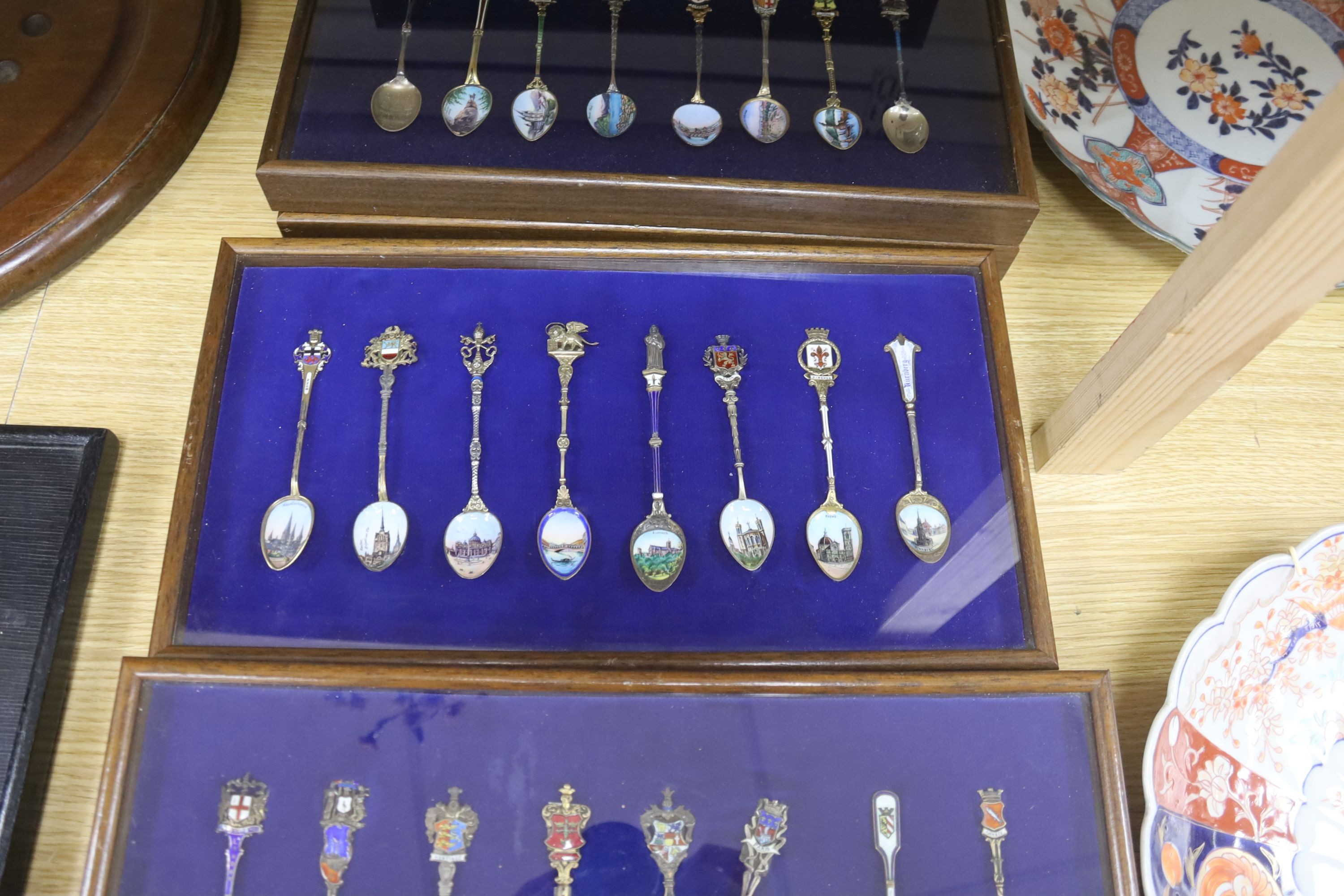 A collection of sixty four 800 standard silver (many gilt) and enamel souvenir spoons, early 20th century, in eight display cases, Including views of London, Germany, Italy, Austria, Spain etc.